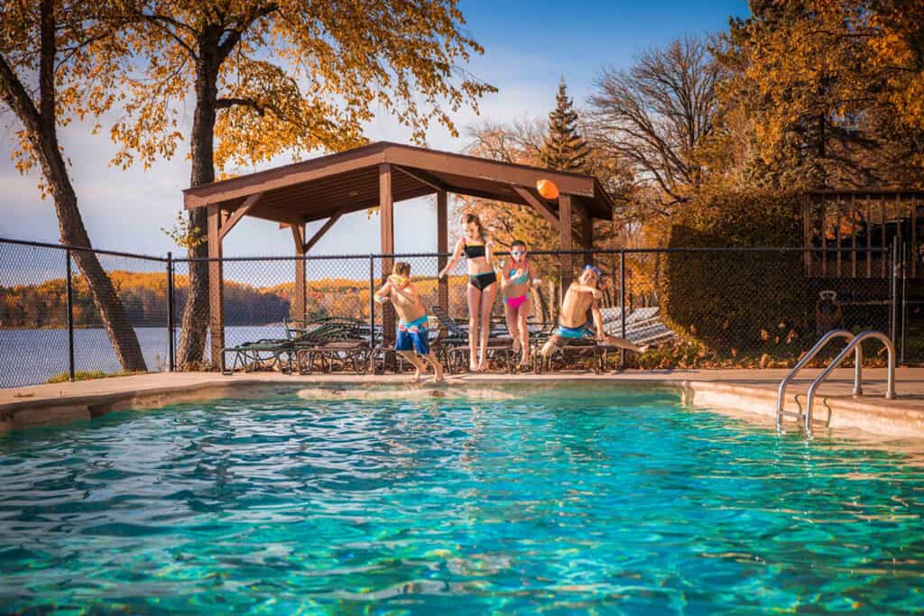 Children jumping into an outdoor pool on a sunny fall day with orange trees and a blue lake in the background at Ruttger's Bay lake Resort