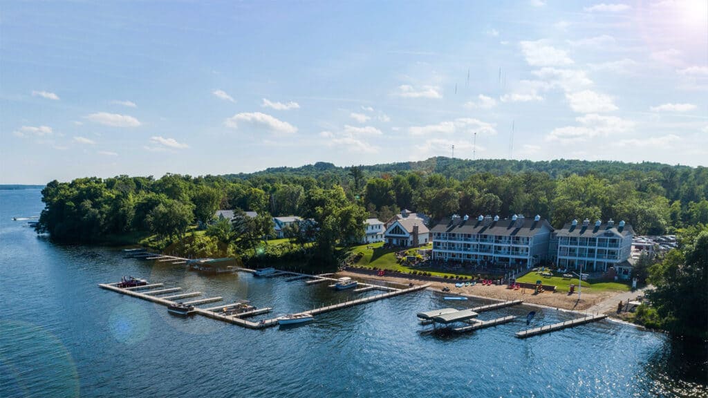 1920x1080 aerial photo of Quarterdeck Resort showing the large three story lodging building along the lakeshore, with a sandy beach and boat docks, and Gull Lake in the foreground and Forest in the Background on a warm sunny summer day