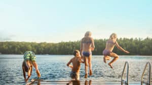1920x1080 photo taken behind kids jumping off of dock into a lake on a warm summer day with the lake and pine trees in the background