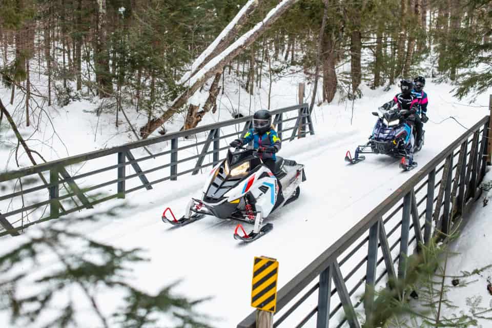 960x640 Photo of two people snowmobiling on a trail in the woods on a bright winter day