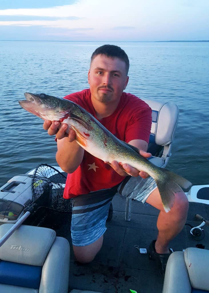 Man holding a walleye in a boat that he just caught with a lake at sunset in the background copy