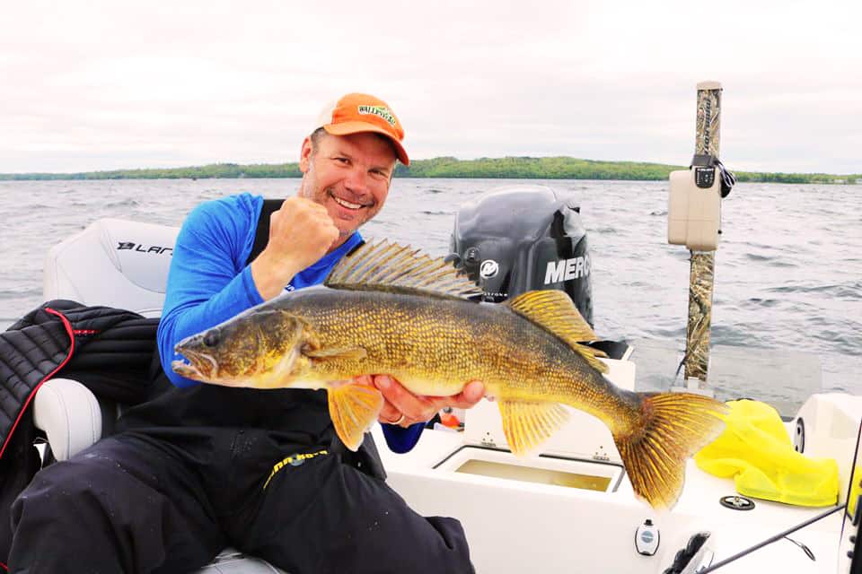 Photo of man holding a walleye in a boat with a lake and shoreline in the background