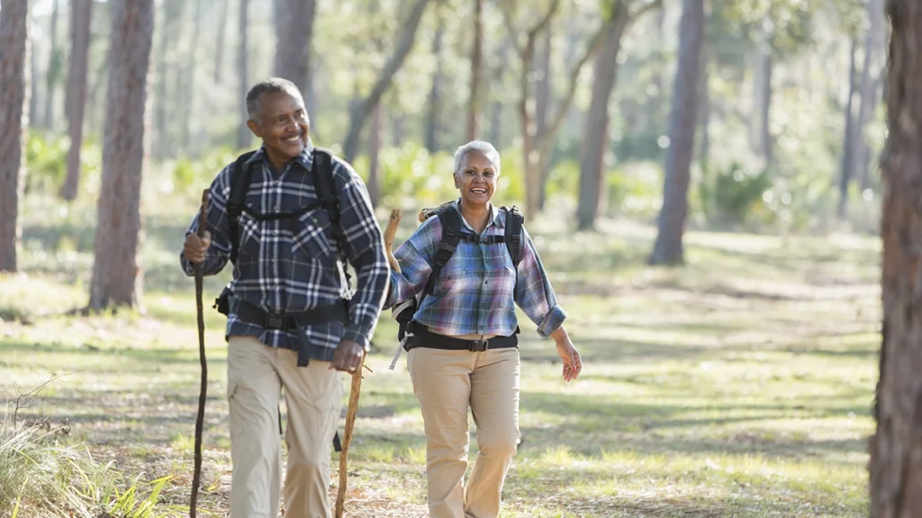 Photo of a smiling older couple wearing backpacks and holding walking sticks taking a hike throught the forest
