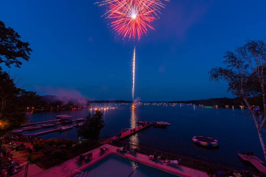 Fireworks high in the sky during dusk over a lake with boats in the water in the distance with a dock and shore in the foreground copy