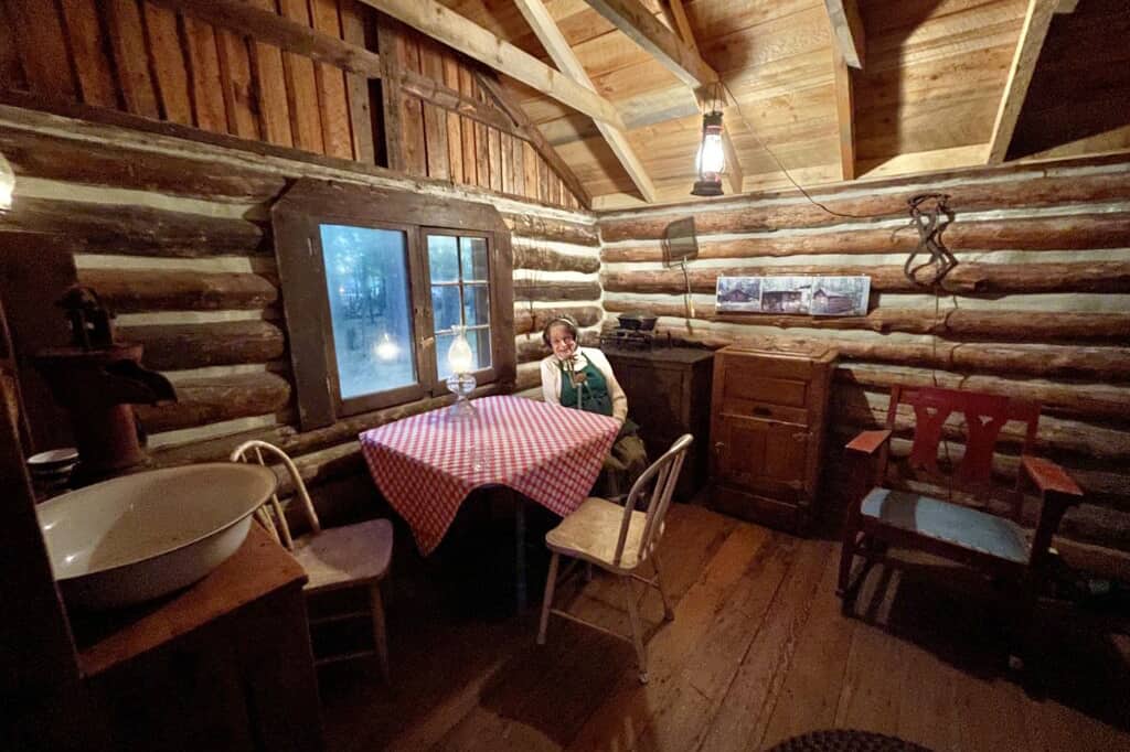 Photo of the interrior of a historical log cabin with a woman dressed up in pionerr era clothing