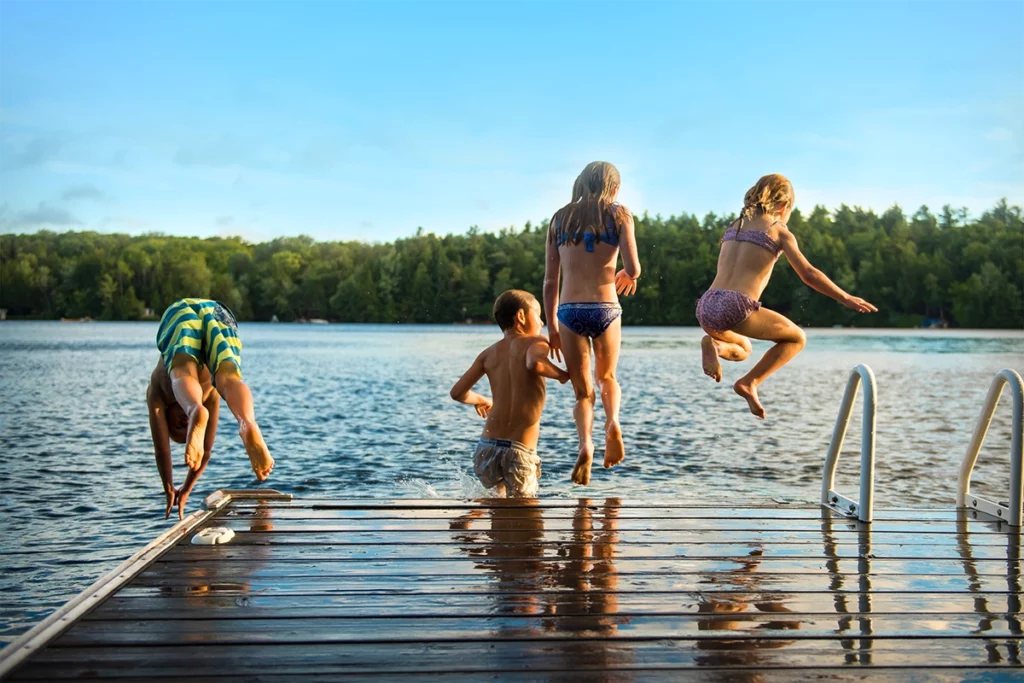 photo taken behind kids jumping off of dock into a lake on a warm summer day with the lake and pine trees in the background