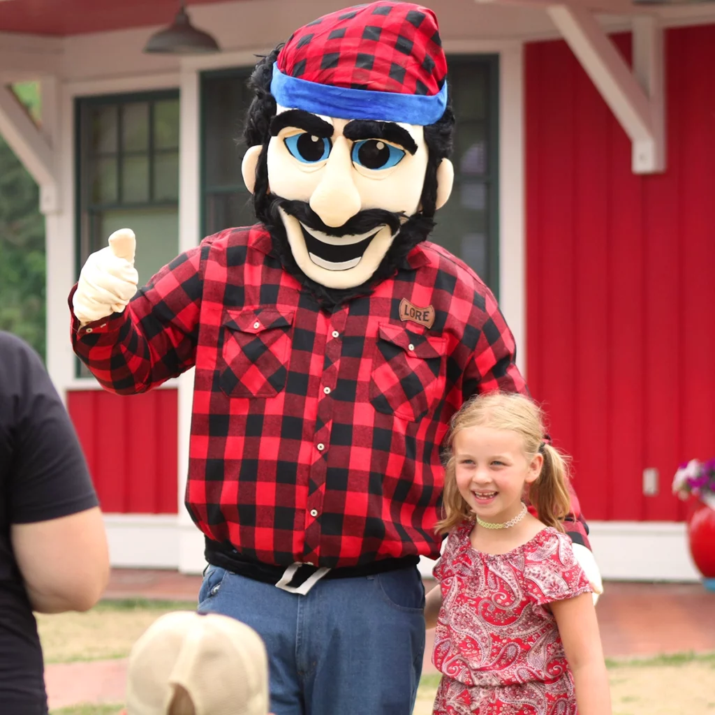 Photo of Bean Hole Days showing a person wearing a Paul Bunayn lumberjack costume giving a thumbs up next to a small, smiling girl with the red Pequot Lakes Chamber building in the background