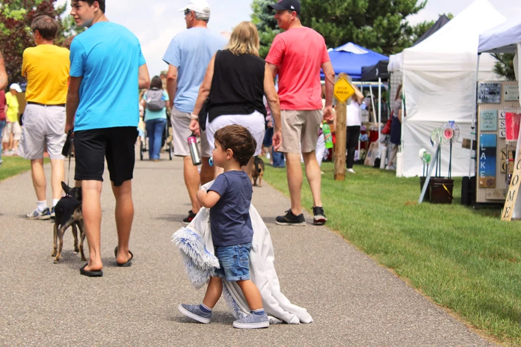 Photo of Bean Hole Days showing a small boy carrying his blanket and walking through the craft fair tents on a path on a summer day with crowds of people in the background