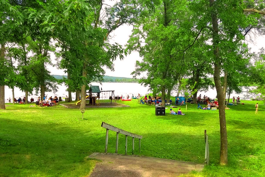 Photo of a park filled with people on a summer day next to a lake with trees and a playground.