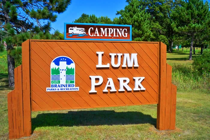 Photo of a sign that says Lum Park Camping on a summer day with trees in the background.