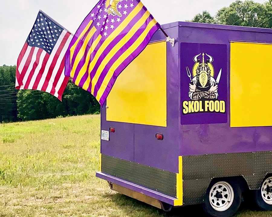 Photo of a yellow and purple food truck with an American Flag and a Minnesota Viking Flag parked on a lawn on a summer day copy