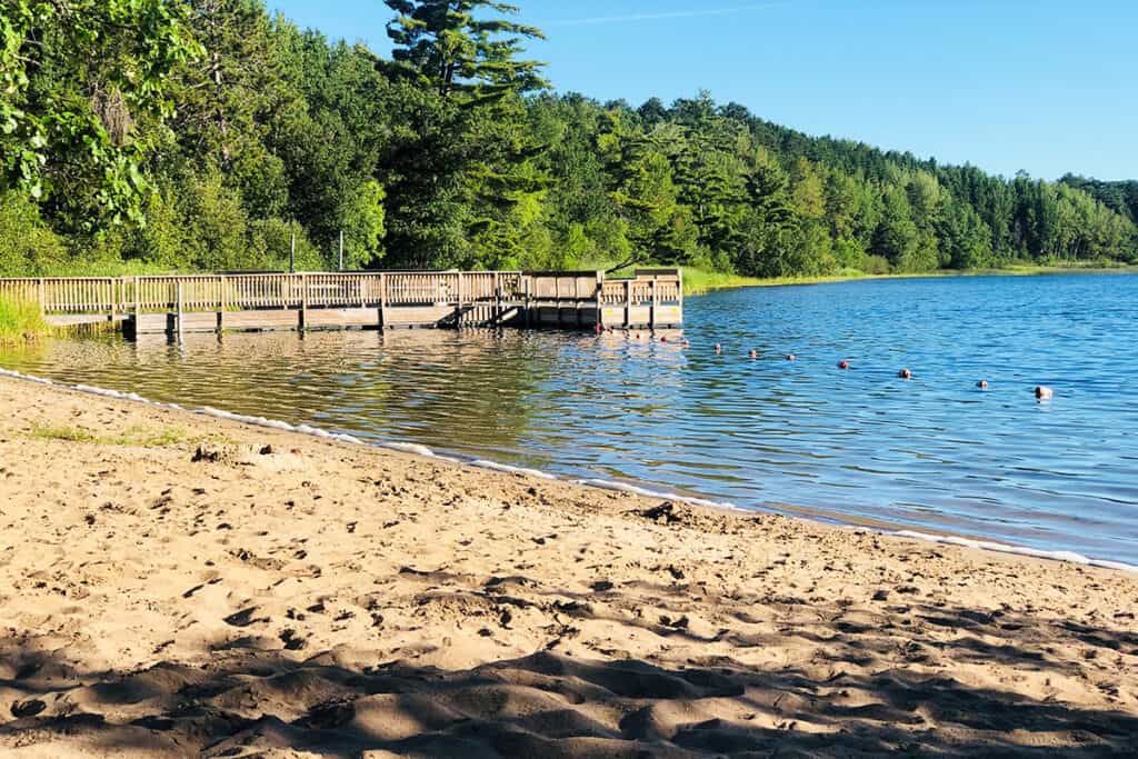 Sandy beach next to a lake with a fishing peir in the background and the lake surrounded by forest on a sunny summer day