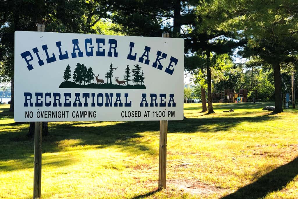 Sign that says Pillager Lake Recreation Area with a wooded park and playground in the background on a sunny summer day