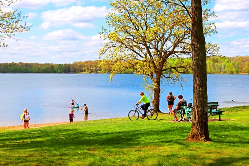 Photo of people swimming and biking at Whipple Beach with a lawn in the foreground and lake in the background on an early fall day.