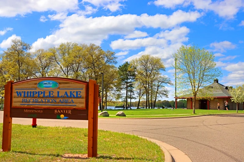 Photo of Whipple Lake Recreation Area sign with a park and lake in the background on a sunny fall day.