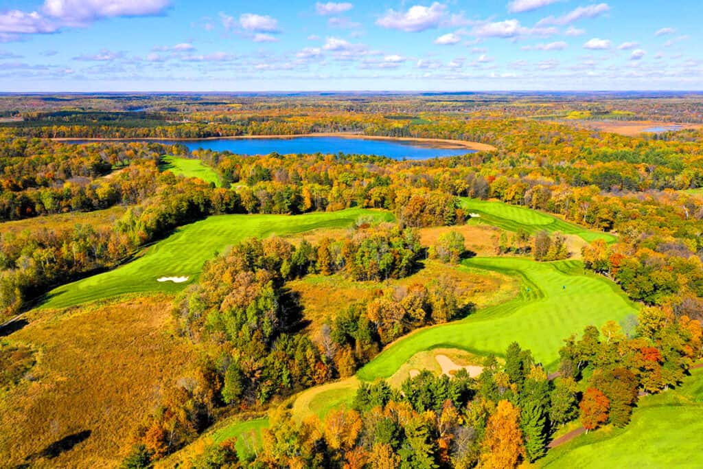 Aerial photo of Deacons Lodge Golf Course at Breezy Point Resort showing the large green golf course surrounded by orange and yellow trees on a sunny fall day with a lake in the background
