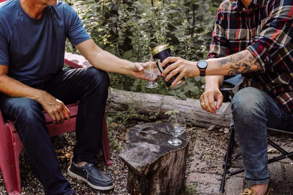 Photo of two young men sharing a cider beer while siting outside on a fall day at a campsite with woods in the background
