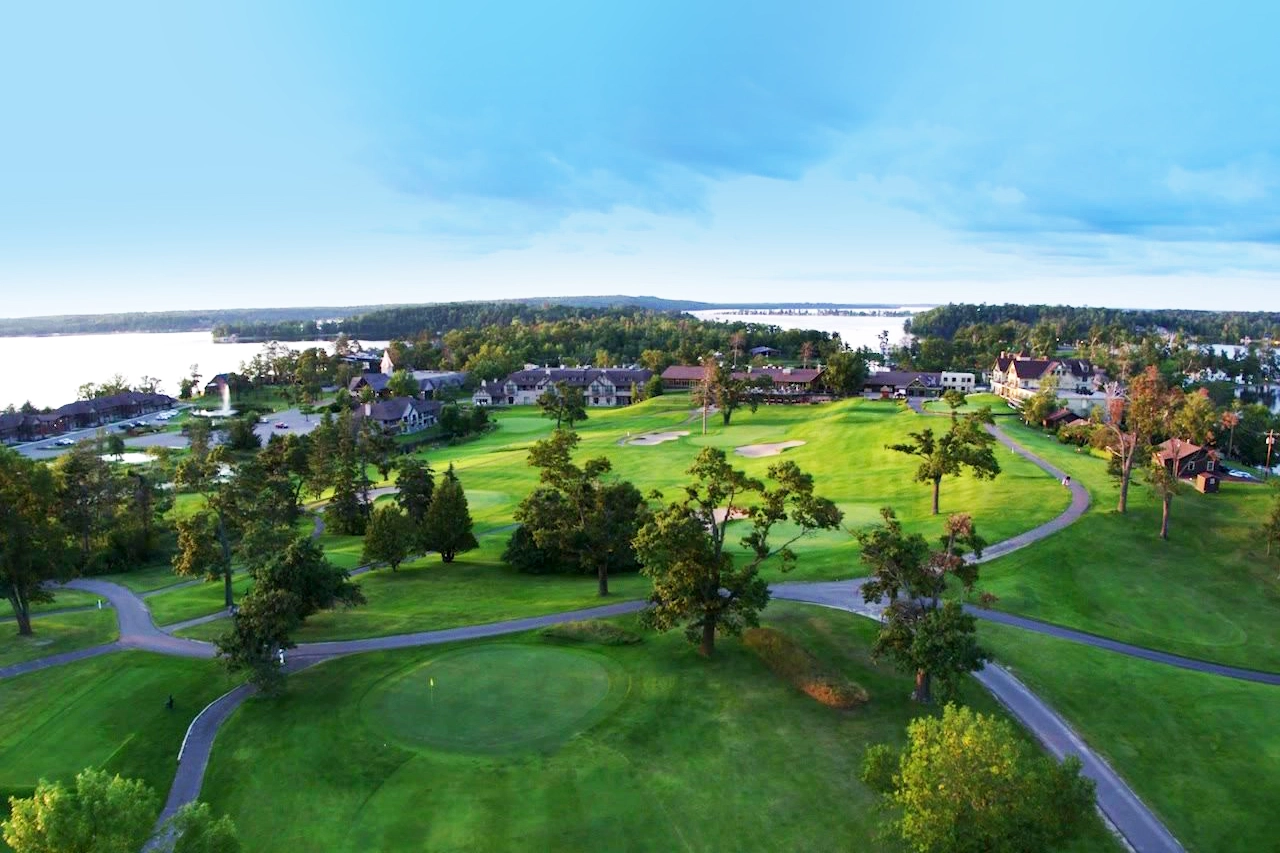 Aerial photo of Maddens on Gull Lake on a summer day showing the vast golf courses with trees and walking paths with the large resort buildings and cabins in the distance and Gull Lake in the background