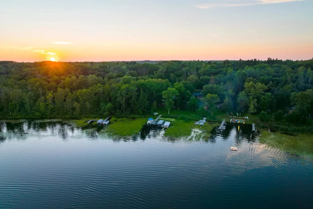 Aerial photo of Tri-Birches Resort showing a hsandy beach at the edge of a lake with large docks boats at the edge of a big woods at sunset on a summer day