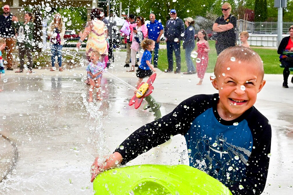 Photo of a young boy playing at the Brainerd Splash Pad at Memorial Park on a summer day with other kids playing in the background and parents watching from the edge of the splash pad