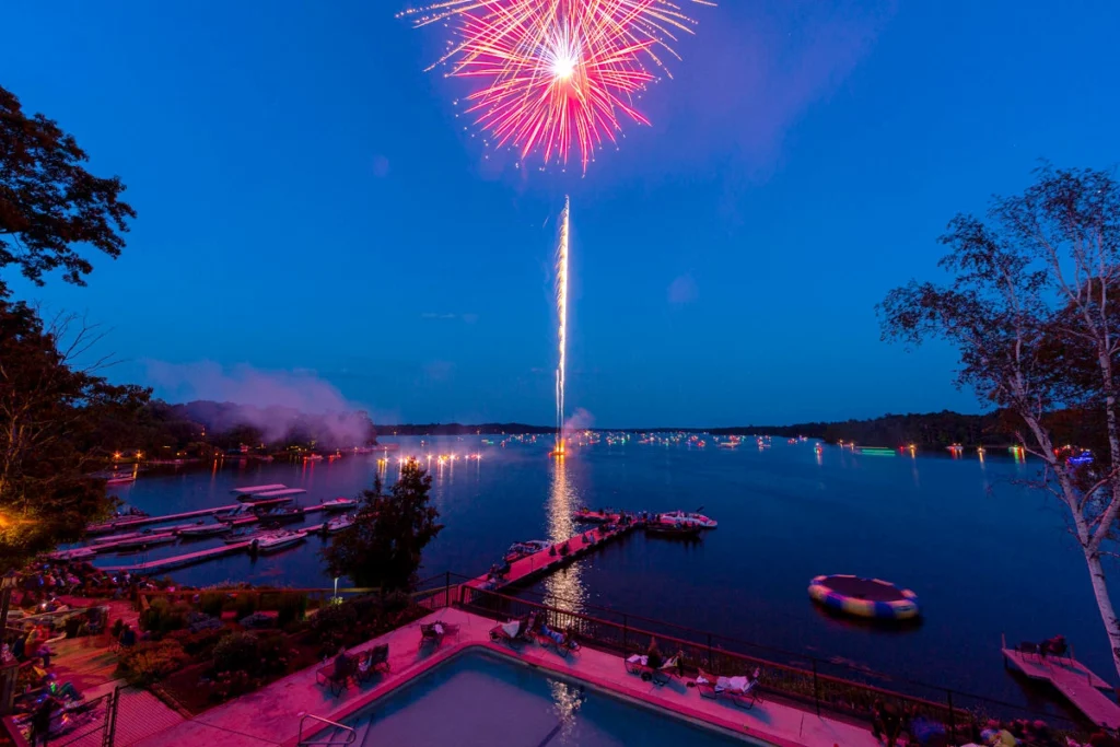 Fireworks high in the sky during dusk over a lake with boats in the water in the distance with a dock and shore in the foreground copy