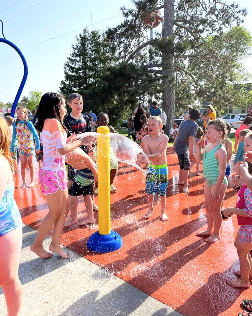 Photo of a large group of young kids playing at the Pequot Lakes Splash Pad in Trailside Park on a summer day with parents watching from the edge of the splash pad