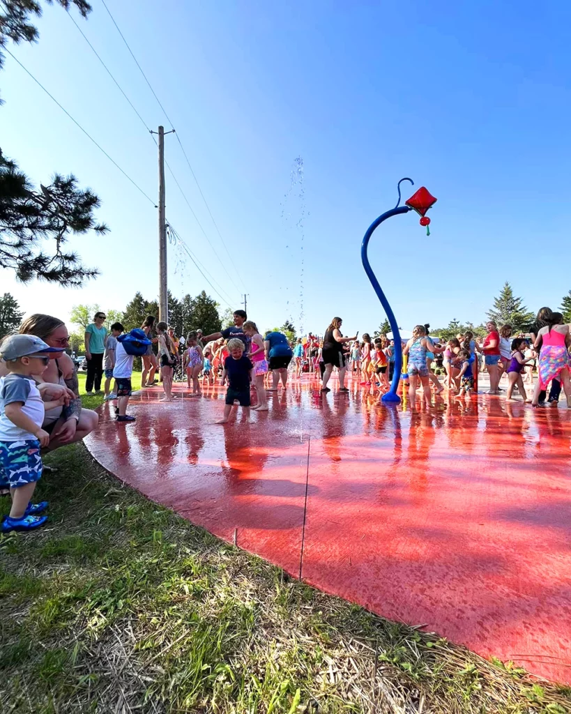 Photo of a large group of young kids playing at the Pequot Lakes Splash Pad in Trailside Park on a summer day with parents watching from the edge of the splash pad