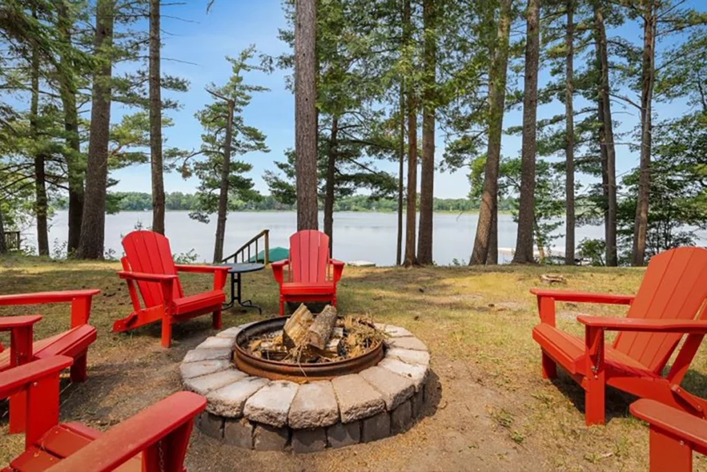 Photo from Book the BLA of Cozy Cabin on Clark Lake showing red lawn chairs sourounding a firepit located near huge pines at the edge of a lake on a summer day copy