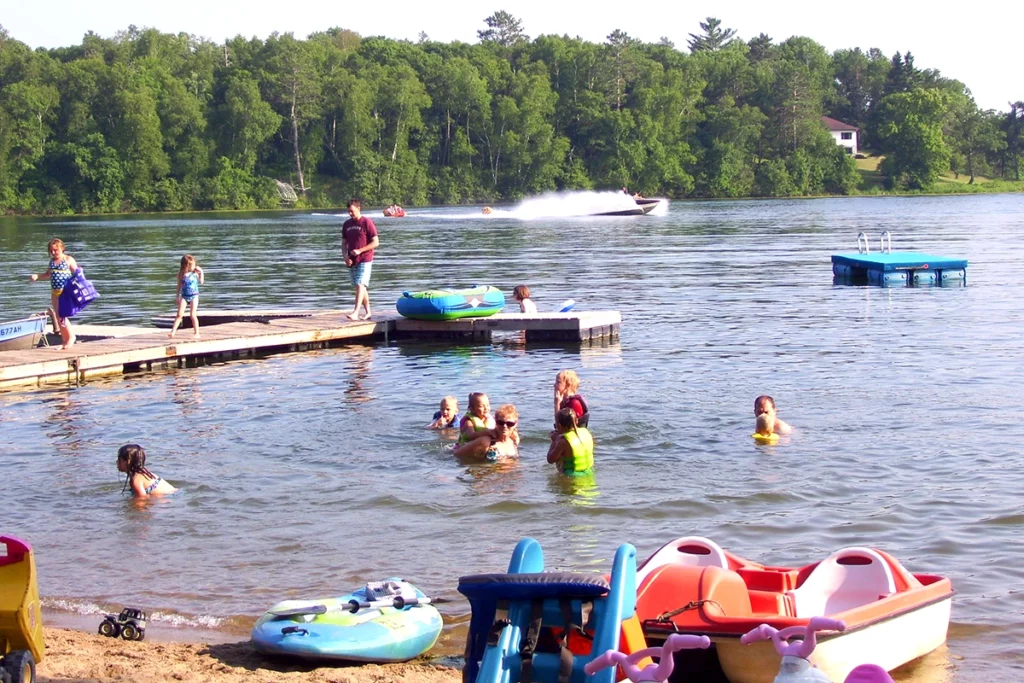 Photo from Shady Hollow Resort showing busy swimming beach with water toys and dock and many kids swimming and playing in the lake on a summer day