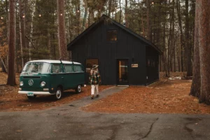 Photo of a black cabin at Nature Link Resort surrounded by large pine trees showing a woman walking into cabin with a vintage van out front