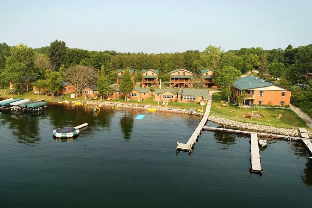 Photo of a classic cabin-style resort with forest in the background and next to a large lake showing docks out front in the water on a sunny summer day