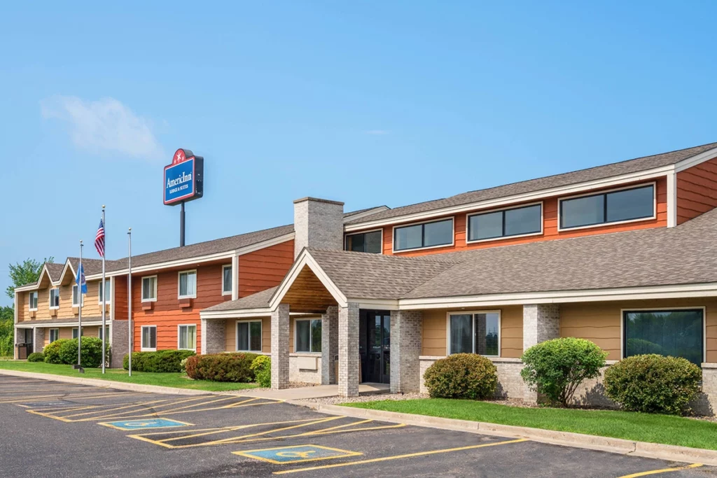 Photo of the exterior of the AmericInn in Little Falls showing mid-size hotel painted brown and orange on a sunny summer day with a blue sky copy