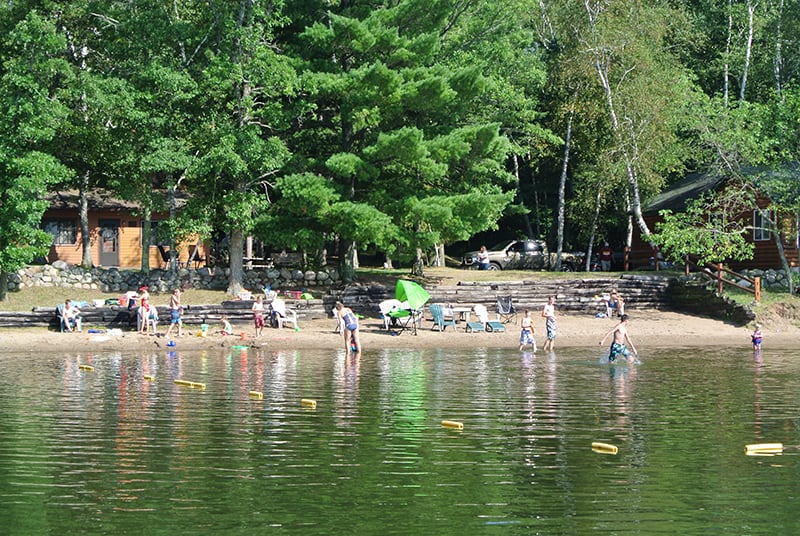 Photo of Galles' Upper Cullen Resort showing a swim beach at the edge of the lake on a summer day with kids playing in the water and woods and cabins in the background