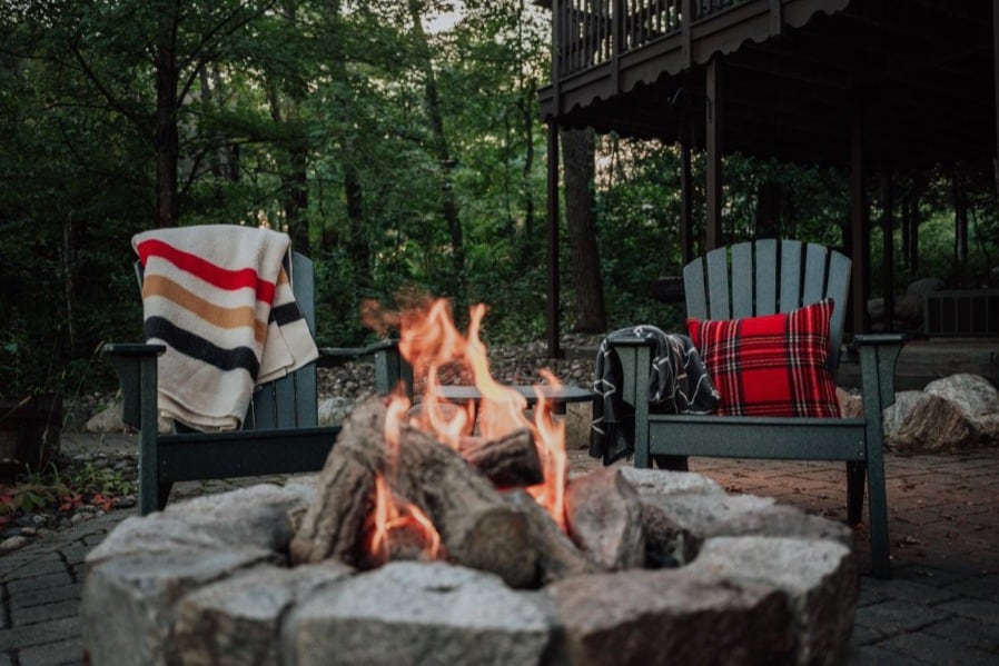 A rustic cabin with woods in the background and a bonfire in the foreground with green Adirondack chairs nestled on Kimble Lake part of the Ossawinnamakee Chain