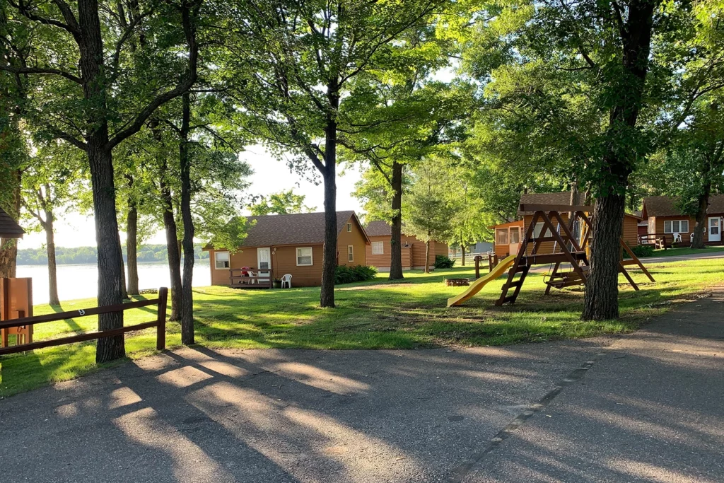 Photo of Train Bell Resort showing several small log cabins at the edge of a lake with big trees and green lawn and a swing set copy