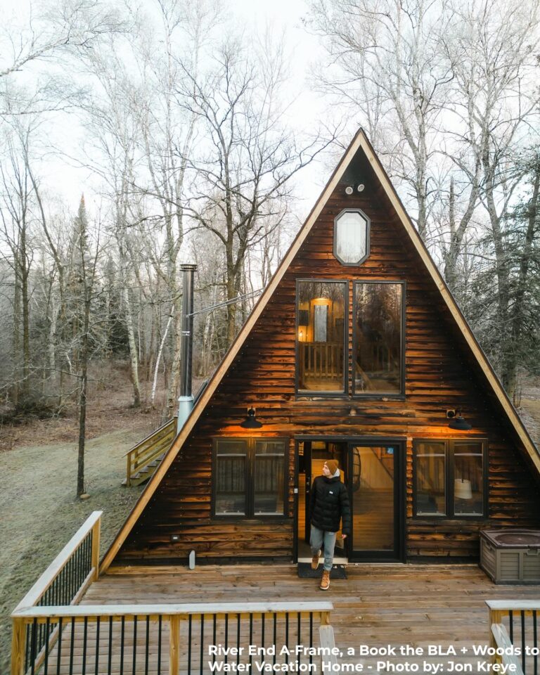 A cozy, dark wood A-frame cabin with large windows and warm interior lighting sits in a clearing surrounded by bare trees. A person in a black coat, tan beanie, and boots steps out onto the spacious wooden deck. The scene has a peaceful, early winter atmosphere. Text at the bottom reads: “River End A-Frame, a Book the BLA + Woods to Water Vacation Home - Photo by: Jon Kreye