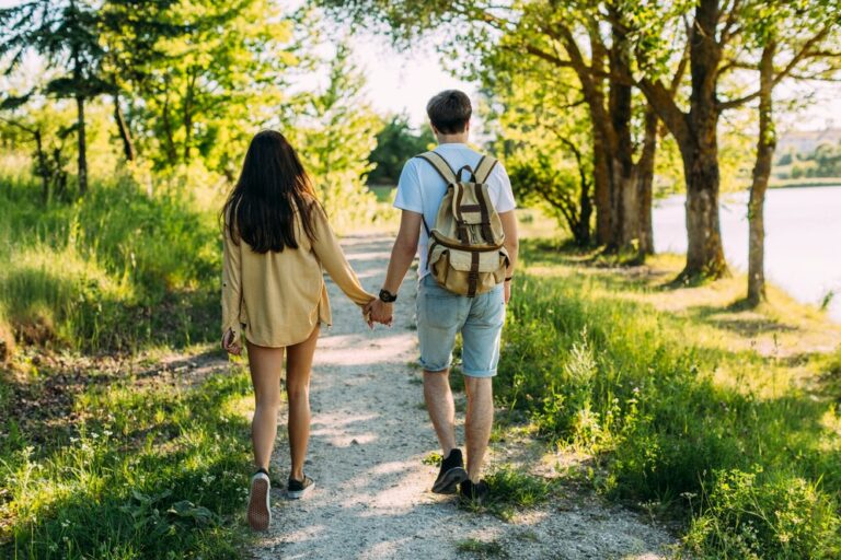 A young couple walks hand in hand down a sunlit gravel path surrounded by trees and greenery. The man wears a backpack and shorts, and the woman is dressed casually in a long-sleeve shirt. A calm body of water is visible to the right, creating a peaceful, nature-filled setting.