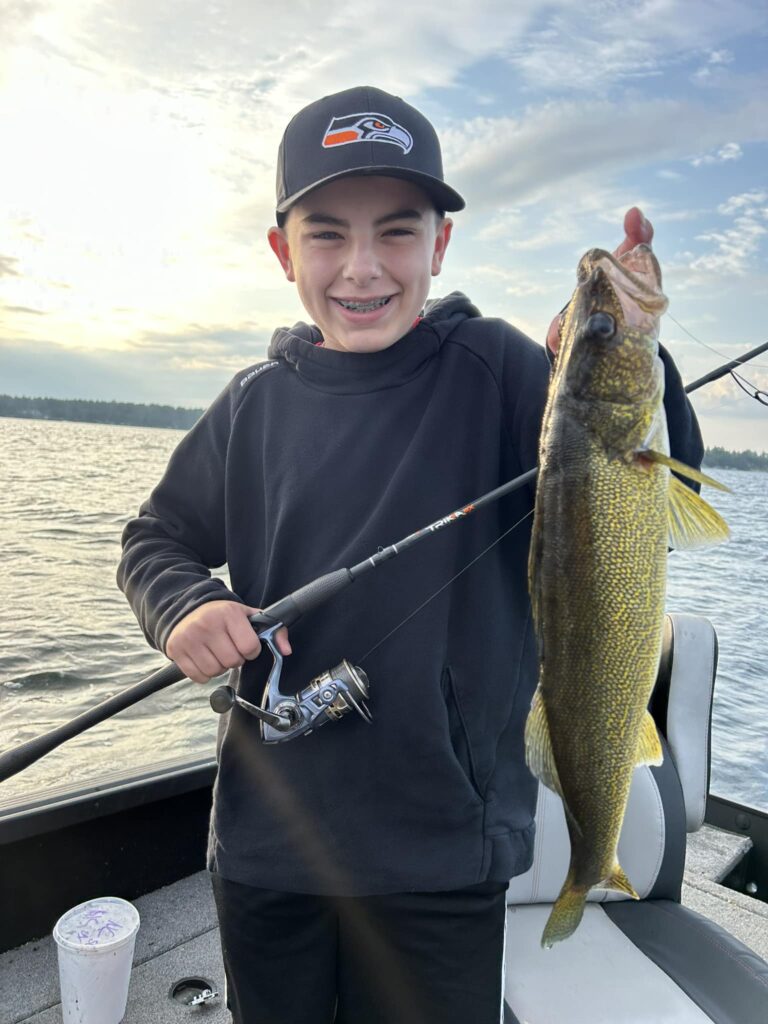 A smiling boy wearing a black hoodie and a Seattle Seahawks hat proudly holds up a freshly caught walleye while standing on a boat at sunrise or sunset on a lake. He grips a fishing rod in his other hand, with the water and distant treeline visible in the background.