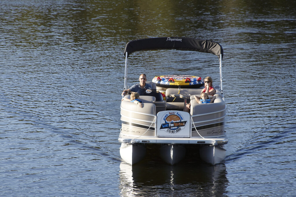 A family rides on a tan pontoon boat labeled "Lake Fun Rentals" across calm lake water, with a colorful inflatable tube on board and two young children wearing life jackets.