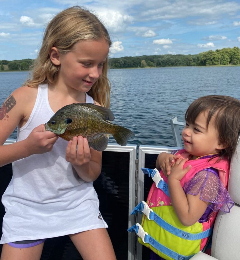 Young girl holding a bluegill fish on a boat shows it to a smiling toddler in a colorful life jacket, with a calm lake and tree-lined shore in the background.