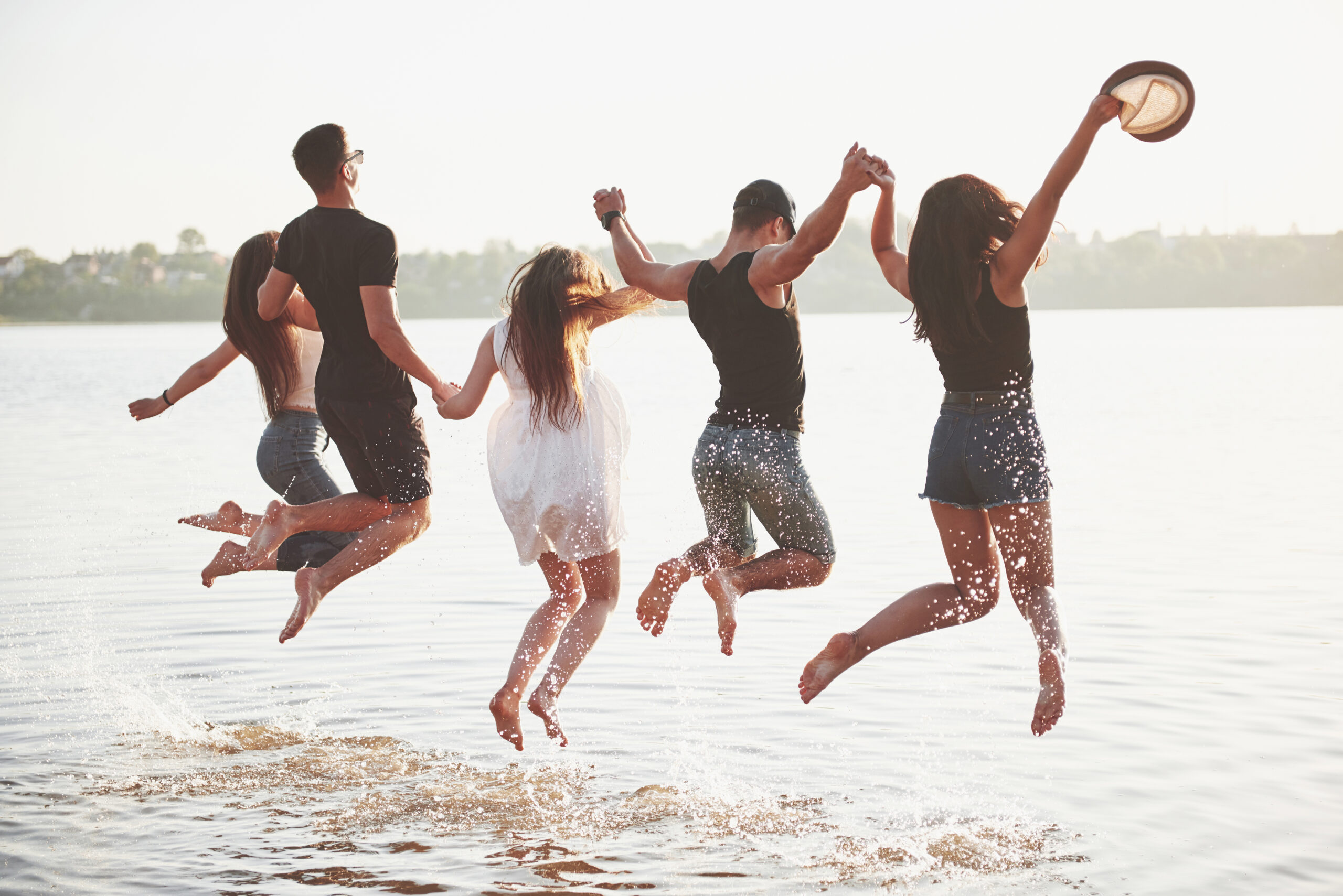 Group of five young adults holding hands and jumping into a lake, splashing water on a sunny day, with a scenic shoreline in the background.